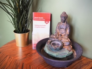 Photo of a Buddha ornament next to a small potted plant with long, tall green leaves, on a wooden table top.