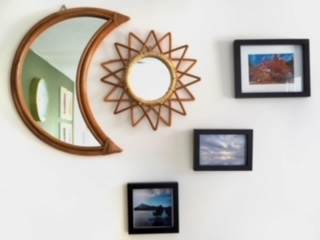 Photo of a half moon shaped mirror, a sun shaped mirror and 3 small scenic photographs hanging on a white wall.